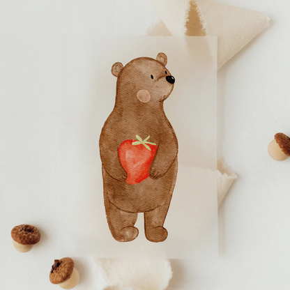 Hier sieht man auf diesem Bügelmotiv eine Bären mit Erdbeere in den Händen. Er schaut zur Seite und hat Pauschbäckchen. Es handelt sich um ein Kindermotiv.