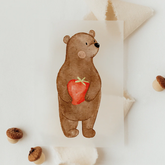 Hier sieht man auf diesem Bügelmotiv eine Bären mit Erdbeere in den Händen. Er schaut zur Seite und hat Pauschbäckchen. Es handelt sich um ein Kindermotiv.