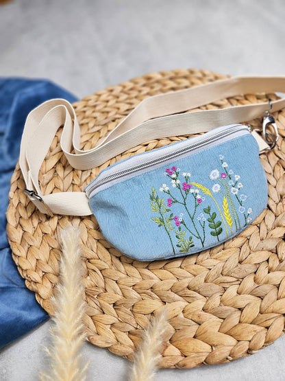Eine hellblaue Bauchtasche aus Cord und schöner Stickerei mit Wildblumenwiese. Der Cord besteht as 100% Biobaumwolle. Die Tasche hat einen weiß silbernen Reißverschluss und ein remefarbenes Gurtband aus 100% Baumwolle. 