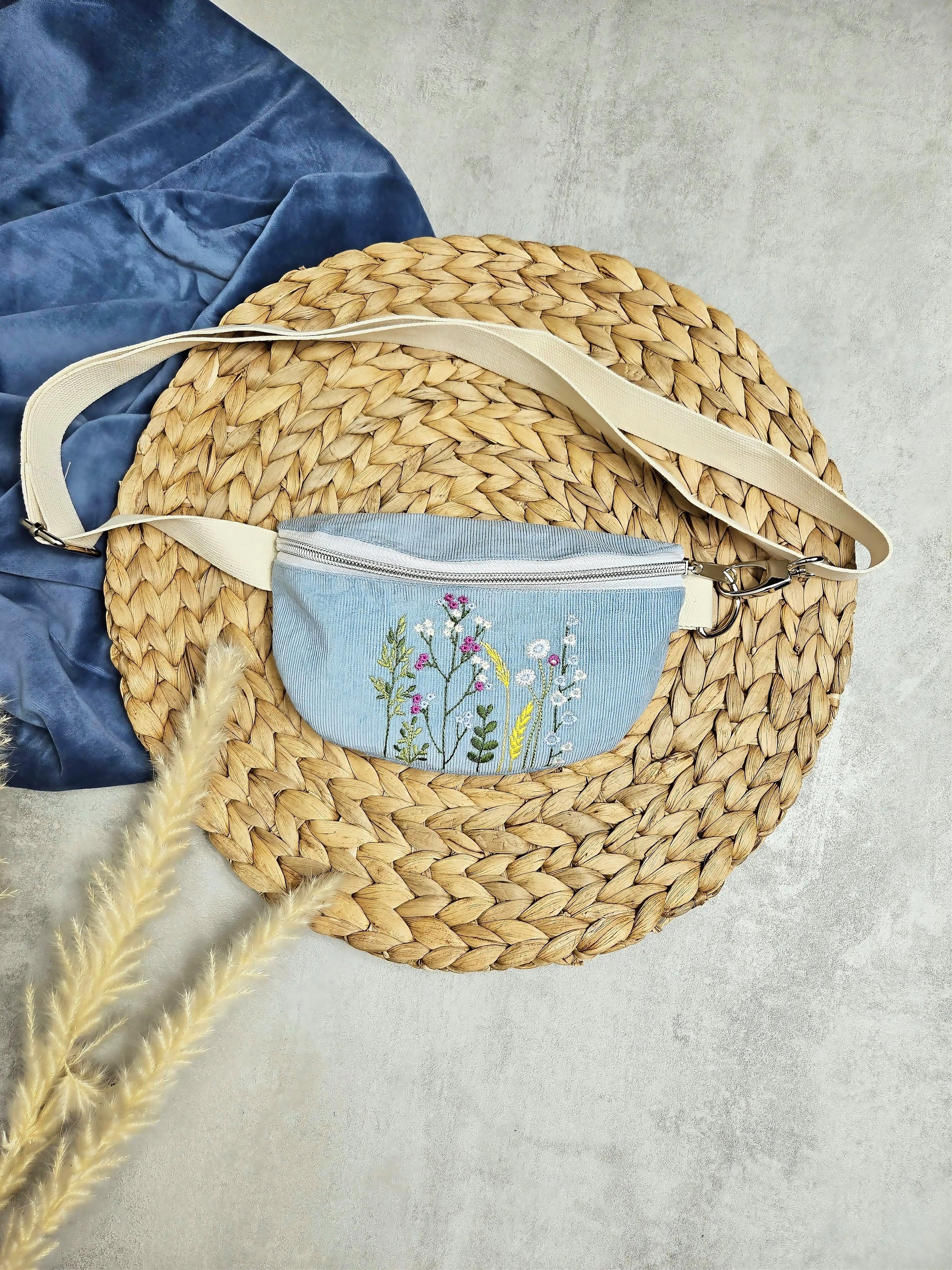 Hier sieht man in einer Frontansicht von oben, die hellblaue Feincord- Umhängetasche mit Wildblumenstickerei. Die Tasche hat silberfarbene Metalldetails und ein helles Gurtband und Reißverschluss.