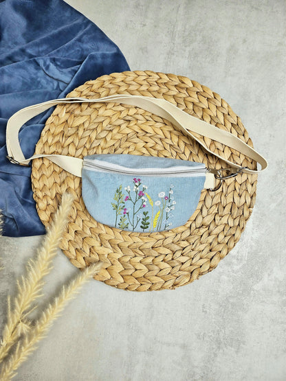 Hier sieht man in einer Frontansicht von oben, die hellblaue Feincord- Umhängetasche mit Wildblumenstickerei. Die Tasche hat silberfarbene Metalldetails und ein helles Gurtband und Reißverschluss.
