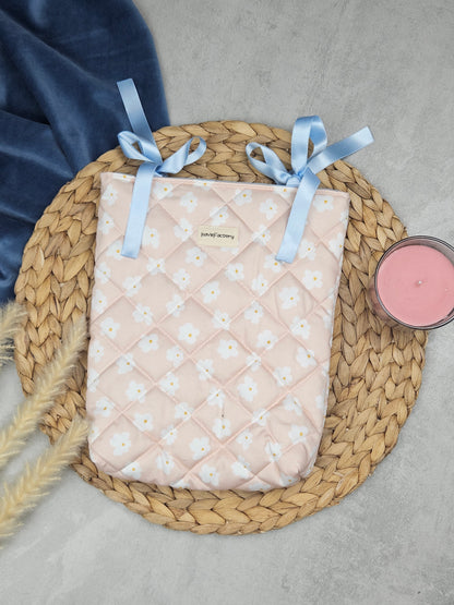 Hier sieht man eine sehr feminine Buchhülle in Rosa mit weißen Gänseblümchen. Verschlossen werden kann die Buchhülle mit hellblauen Schleifen. Die Hülle ist wattiert mit einem 100% recyceltem Volumenvlies und von außen diagonal abgesteppt.
