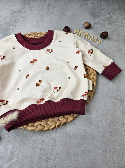 Heller Kindersweater mit dezentem Pilzmotiv und weinroten Bündchen